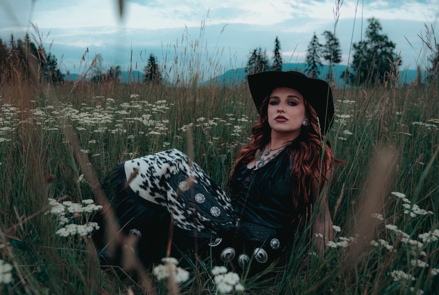 A woman with auburn hair wearing a black dress and cowboy hat sat in a field